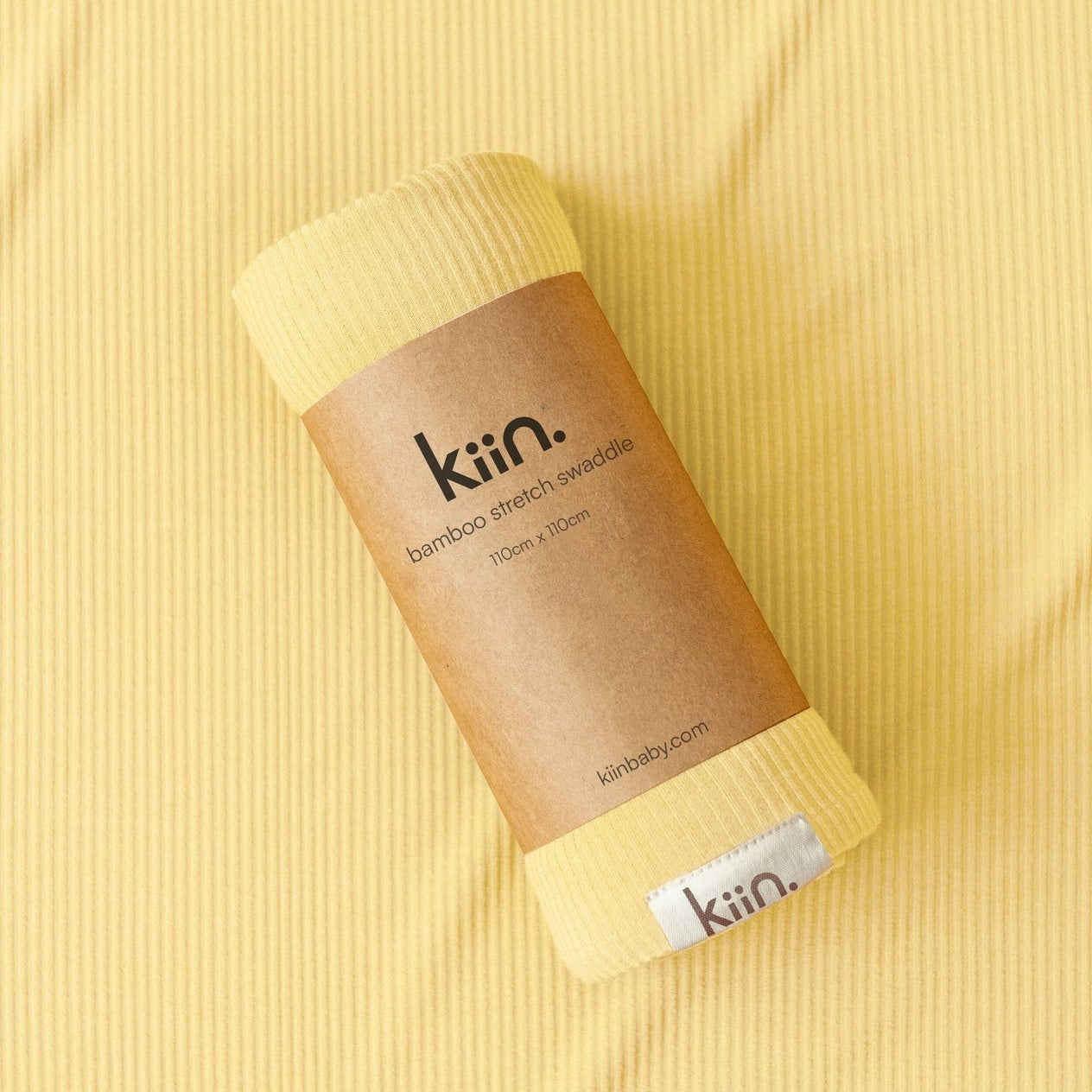 A rolled bamboo stretch swaddle blanket in pale yellow, wrapped in a brown kraft paper band labeled 'kin.', placed on a matching ribbed yellow fabric background.