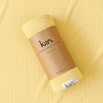 A rolled bamboo stretch swaddle blanket in pale yellow, wrapped in a brown kraft paper band labeled 'kin.', placed on a matching ribbed yellow fabric background.