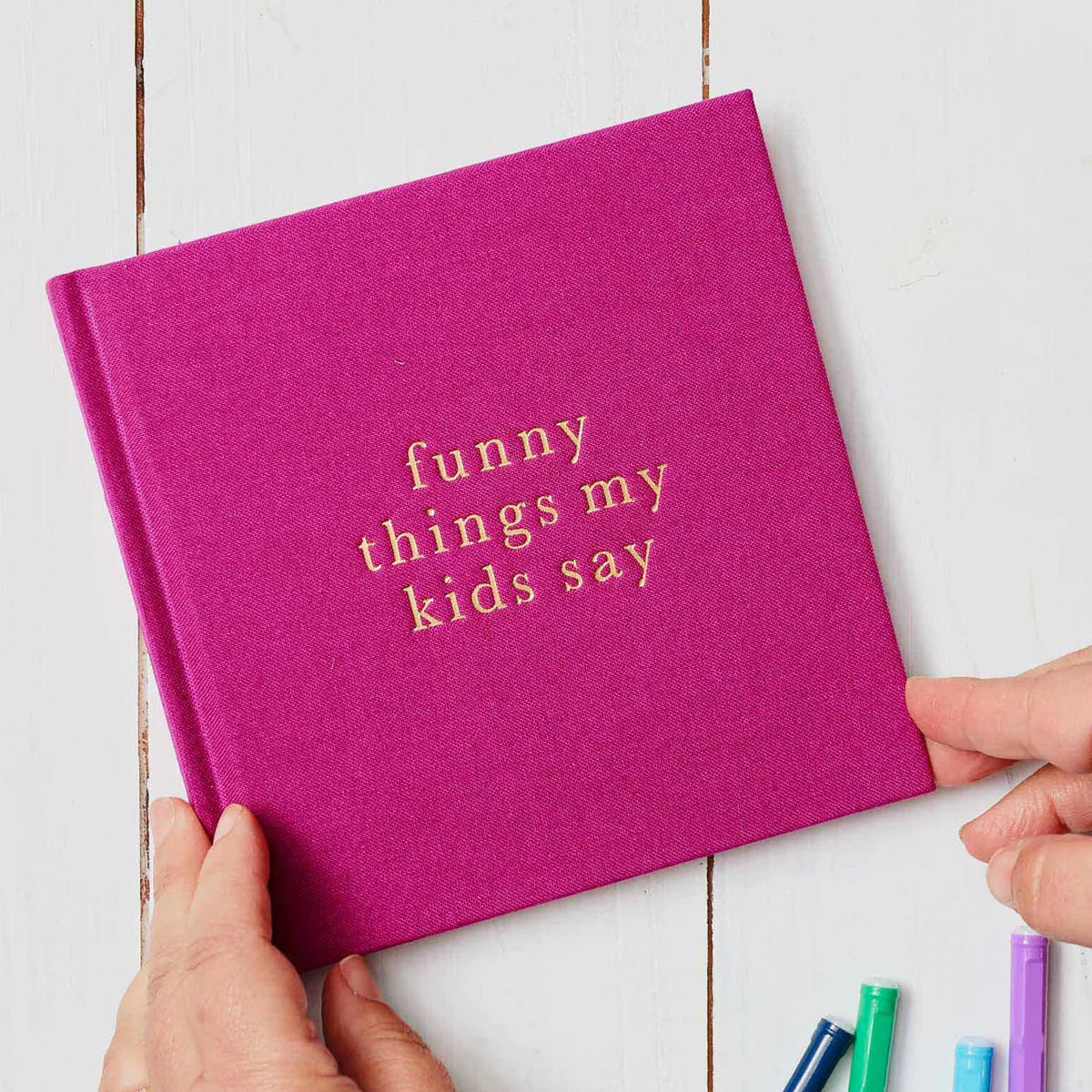 Pink book titled 'funny things my kids say' held by hands on a white wooden surface with colorful pens.